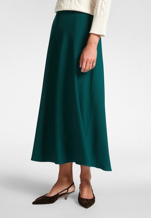 Falda midi verde y fluida, hecha de una tela suave, con una cinturilla ancha y un sutil drapeado, combinada con tacones slingback marrones de punta.