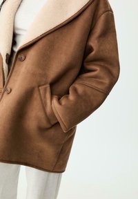 Hnedá umelá zamšová bunda s krémovým ovčím golierom, zapínaním na gombiky a bočnými vreckami. Mäkká textúra a nadmerný strih sú výrazné vlastnosti.