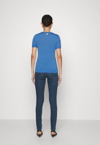Mujer con un peinado de moño oscuro, vistiendo una blusa azul de mangas cortas, jeans oscuros ajustados y sandalias de tacón negras, de pie frente a una pared blanca.