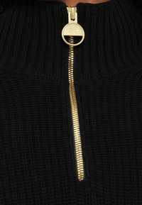 Fekete kötött pulóver magas gallérral, arany cipzárral díszítve. A textúra bordázott, ami kiemeli a ruhadarab részleteit.