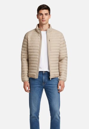 Giovane uomo con giacca trapuntata beige sopra una t-shirt bianca e jeans blu, in piedi, rivolto verso la fotocamera con espressione neutra.