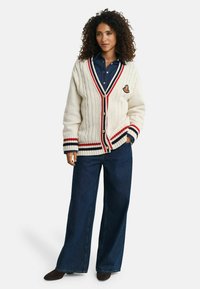 Frau mit lockigem Haar, die eine crèmefarbene Strickjacke mit roten, weißen und maritimen Streifen über einem dunkelblauen Denimhemd und weit geschnittenen Jeans trägt.