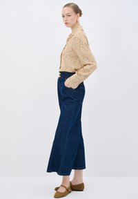 Camisa de botones beige con estampado, jeans de pierna ancha de cintura alta en azul oscuro y zapatos planos de ante marrón; el modelo posa de lado contra un fondo liso.