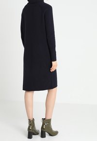 Robe en tricot bleu marine avec un col haut, des manches longues et une coupe droite, associée à des bottes chevilles vert olive avec des talons épais.