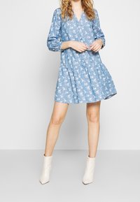 Niebieska sukienka w kwiatowy wzór z dekoltem w kształcie litery V, długimi bufkami, warstwową spódnicą i miękką strukturą. Noszona z beżowymi botkami za kostkę.