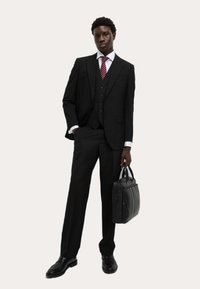 Czarny garnitur trzyczęściowy z białą koszulą i bordową wąską krawatem. Trzyma czarną skórzaną torbę. Polerowane czarne buty. Jednolity tło.