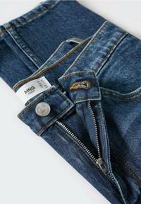 Jean en denim bleu foncé, avec des surpiqûres en cuivre, une fermeture par bouton argenté et une fermeture éclair. Étiquette de marque visible : MNG Denim.