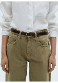 Khakifarbene Baumwollhosen mit lockerer Passform, fünf Taschen-Design und einem braunen Leder-Gürtel mit quadratischer Schnalle. Weißes Hemd mit Knopfleiste.