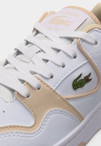 Κοντινό πλάνο λευκού αθλητικού παπουτσιού με μπεζ λεπτομέρειες, λευκά κορδόνια και πράσινο λογότυπο κροκοδείλου στο πλάι, με ραμμένο σχέδιο και επενδυμένη γλώσσα.