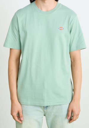 T-shirt en coton vert menthe avec un col rond, des manches courtes, et un petit logo cousu sur le haut gauche de la poitrine.
