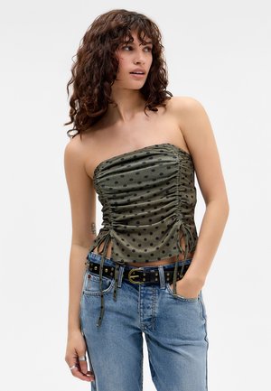 Vrouw met krullend haar die een groene, gerimpelde, strapless polkadot top en blauwe spijkerbroek met een zwarte riem met studs draagt, hand in zak, zijwaarts kijkend.