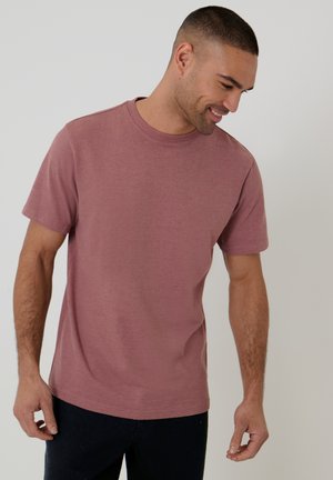 Uomo con t-shirt a maniche corte rosa polveroso e pantaloni neri, guarda verso il basso e sorride su uno sfondo chiaro uniforme.