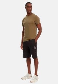 T-shirt verde oliva, design aderente, abbinata a pantaloni corti neri con logo. Il modello indossa sneaker grigio chiaro con accenti beige.