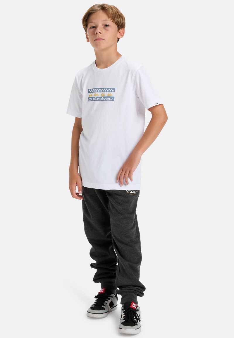 Weißes Baumwoll-T-Shirt mit blauem und gelbem Grafikdesign. Kombiniert mit dunkelgrauen Jogginghosen und schwarzen Turnschuhen mit roten Akzenten.