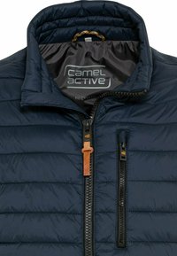 Marineblaue gesteppte Jacke mit zwei vertikalen schwarzen Reißverschlüssen, einer mit braunem Leder-Zuglasche, und hohem Kragen mit Markenlogo innen.
