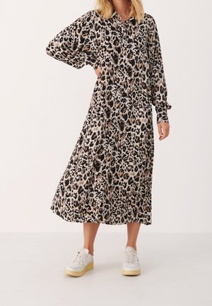 Femme portant une longue robe imprimé léopard avec des boutons et des baskets blanches, debout avec une main sur la hanche.