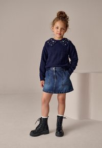 Marineblauer Strickpullover mit einer mit Perlen verzierten Kragen, kombiniert mit einem blauen Jeansrock und schwarzen Schnürstiefeln mit dicken Sohlen.