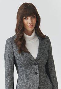 Ušitý šedý blazer s texturovanou látkou, který má klopový límec a dva černé knoflíky. Nosený přes bílý rolák.