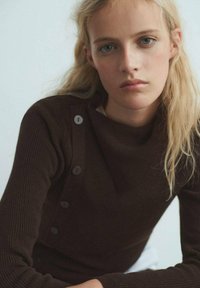 Pull marron c�ôtelé avec un col drapé, présentant quatre grands boutons sur l'épaule gauche. Texture douce, design ajusté.