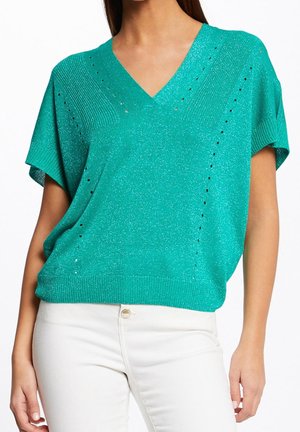 Vrouw die een glinsterende turquoise trui met korte mouwen en V-hals draagt, gecombineerd met witte hoge taille broek, zichtbaar van schouders tot heupen.