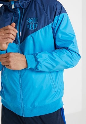 Person, die eine blau-navyfarbene Jacke mit dem FCB-Fußballclublogo auf der Brust zumacht.