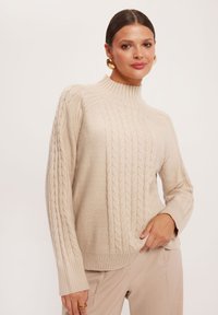 Beżowy sweter w warkocz z wysokim dekoltem i ściągaczami, z teksturowanymi wzorami i nieco luźnym krojem.