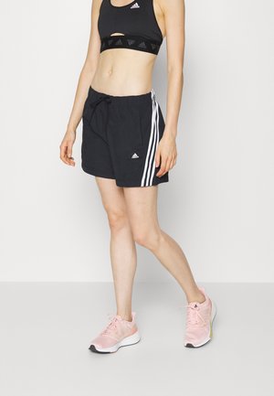 Kobieta w czarnym sportowym staniku i szortach Adidas z białymi paskami, różowe buty do biegania, stoi na białym tle.