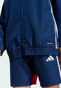 Veste de sport bleu marine avec des rayures blanches, ourlet élastique et logo AeroReady ; associée à un short bleu marine assorti avec une touche de rouge et le logo Adidas.