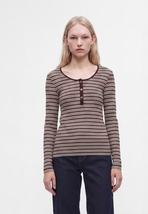 Junge Frau trägt ein braun-weiß gestreiftes Henley-Shirt mit langen Ärmeln und dunkelblaue Jeans, steht vor einem schlichten weißen Hintergrund.