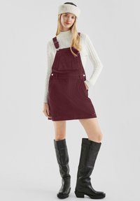 Burgunderfarbiges Cordkleid mit verstellbaren Trägern, Fronttasche, kombiniert mit einem weißen Kabelstrickpullover und schwarzen kniehohen Stiefeln.