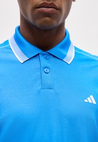 Άνδρας που φοράει φωτεινό μπλε πόλο με γιακά με λευκή περιμετρική ρίγα και δύο κουμπιά κλειστά, με ένα μικρό λευκό λογότυπο στο στήθος.