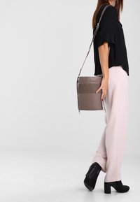Borsa a tracolla grigia in pelle con tracolla regolabile, forma rettangolare, texture liscia e due tasche frontali. Indossata con pantaloni rosa chiaro.