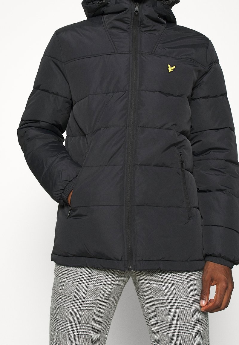 Doudoune noire en tissu durable, avec des sections matelassées, une fermeture éclair, une capuche réglable et un petit logo jaune sur la poitrine.