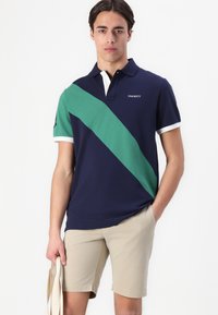 Marineblå polo skjorte med en grønn diagonal stripe, hvit krage og ermekanter. Logoen vises på venstre bryst. Båret med beige shorts.