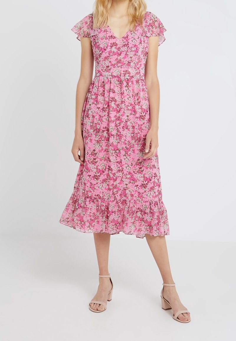 Femme portant une robe midi rose à fleurs avec des manches courtes volantées, un col en V, une jupe froncée et des sandales beige à talons carrés.