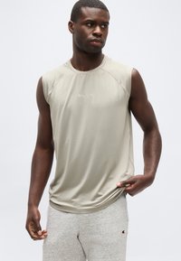 Μυώδης άνδρας με μπεζ αμάνικη μπλούζα Champion και ανοιχτό γκρι φόρμα, διορθώνει το κάτω μέρος της μπλούζας με το δεξί χέρι.