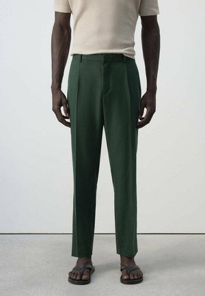 Mann trägt dunkelgrüne maßgeschneiderte Hose, beige kurzärmliges Ripphemd und schwarze Sandalen, steht vor hellem Hintergrund.