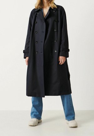 Donna che indossa un lungo trench coat scuro sopra jeans blu e scarpe bianche, in piedi davanti a uno sfondo chiaro uniforme.