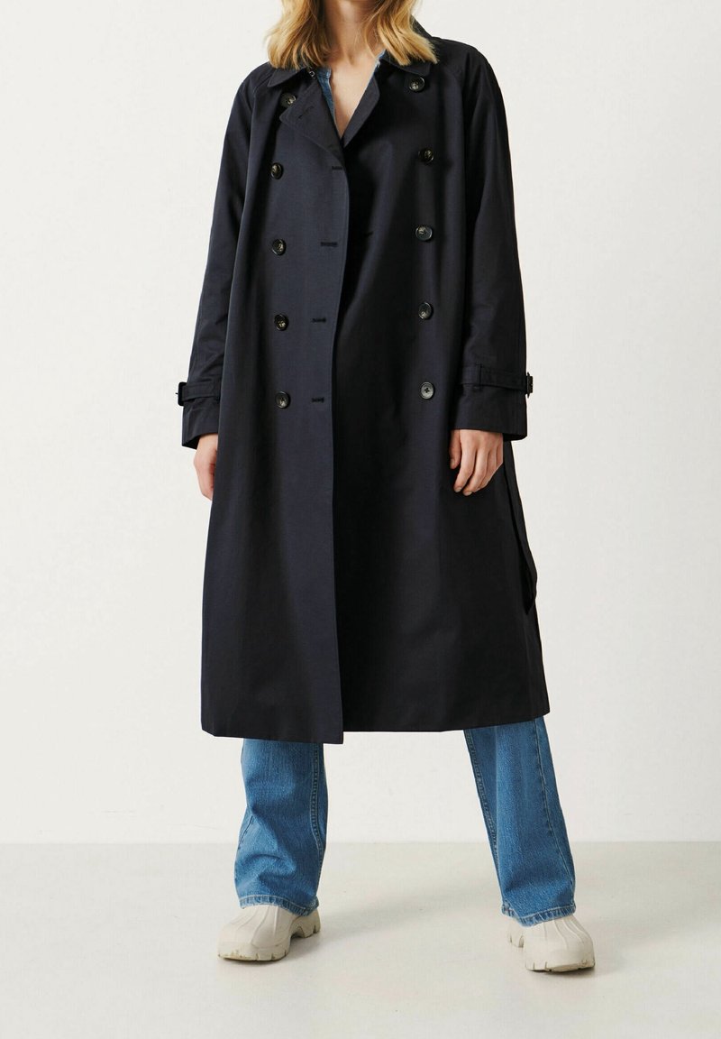 Femme portant un long trench-coat foncé par-dessus un jean bleu et des chaussures blanches, debout devant un fond clair uni.