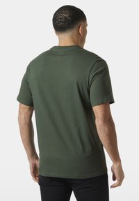 Hombre con cabello rizado corto y pecas, vistiendo una camiseta verde oscura de manga corta lisa y pantalones negros, de espaldas contra un fondo blanco.