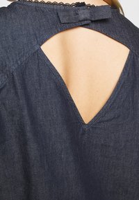 Top in denim con un profondo scollo a V sul retro, con un accento a fiocco e delicate rifiniture in pizzo lungo il collo. Tessuto blu scuro con una consistenza liscia.