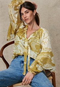 Bluză florală galbenă și albă cu mâneci bufante, decolteu plisat și legare în față, asortată cu blugi din denim de culoare bleu deschis.