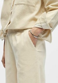 Conjunto de pana color crema con un patrón texturizado, que presenta un ajuste holgado, puños remangados y un bolsillo lateral. También incluye una pulsera metálica.