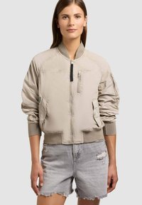 Žena oblečená v béžové bomber bundě se zipovými kapsami a žebrovanými manžetami, kombinované se světle šedými roztrhanými džínovými šortkami.