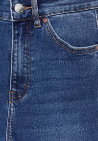 Gros plan sur un jean en denim bleu montrant la poche avant, une couture orange et un bouton argenté avec gravure de la marque.