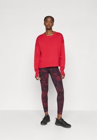 Rød hettegenser med lange ermer og ribbestrikket kant, kombinert med svarte og røde mønstrede leggings. Svarte treningssko fullfører antrekket.