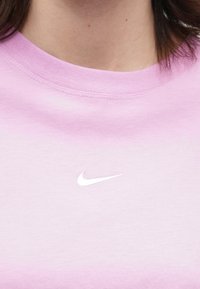 Heled roosa ümar koonuskaelusega särk, millel on valge Nike swooshi logo, mis asub keskelt rinnal, nähtav osa inimesest kaelast ja juustest.