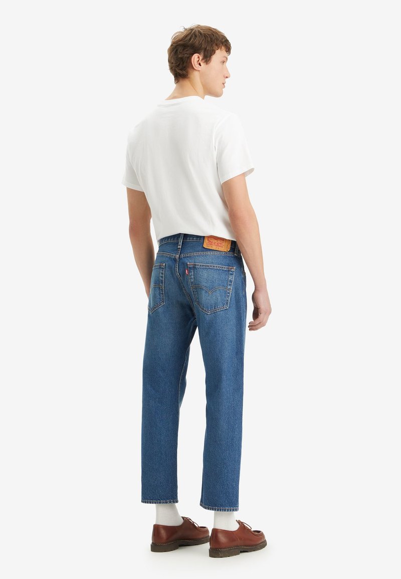 Pantalones vaqueros de mezclilla azul con ajuste recto, con un parche de logo de cuero amarillo en la parte trasera, combinados con una camiseta blanca sencilla y zapatos marrones.