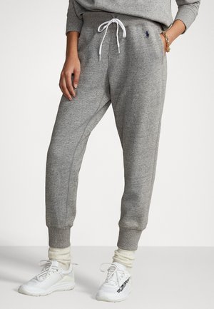 Persoon die grijze joggingbroek draagt met trekkoord en geribbelde boorden, witte sokken, witte sneakers en een gouden armband aan één pols.