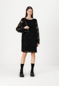 Svart spetklänning med långa ärmar, geometriska mönster och rak skärning. Bärs med en svart handväska och skor med tjock sula.